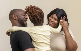 A Black man holding a young Black child whose facing away from the camera on their hip while standing close to a Black woman, with all three positioned indoors against a plain light‑coloured wall. The child’s arms stretch out toward both adults as the group stands closely together.