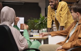 A diverse group of colleagues sit together in an office, smiling and talking. A man in a yellow jacket leans on a desk while two women—one wearing a hijab—sit nearby holding drinks, engaged in friendly conversation.